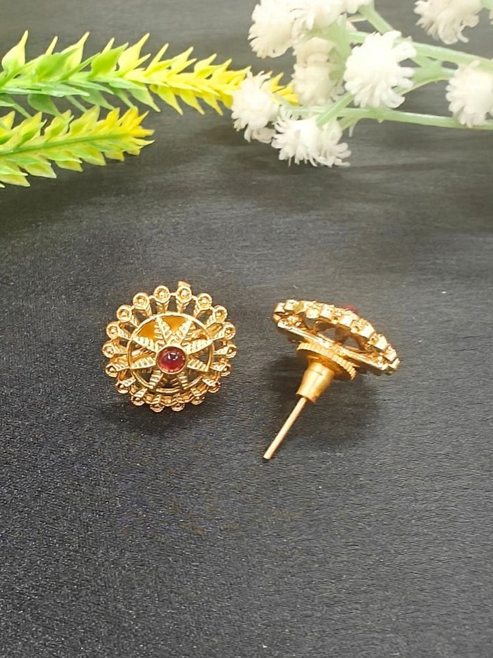 The Red Gehna Earrings are traditional round gold stud earrings with intricate designs and a central ruby-colored stone, displayed on a dark surface with white and yellow artificial flowers in the background.