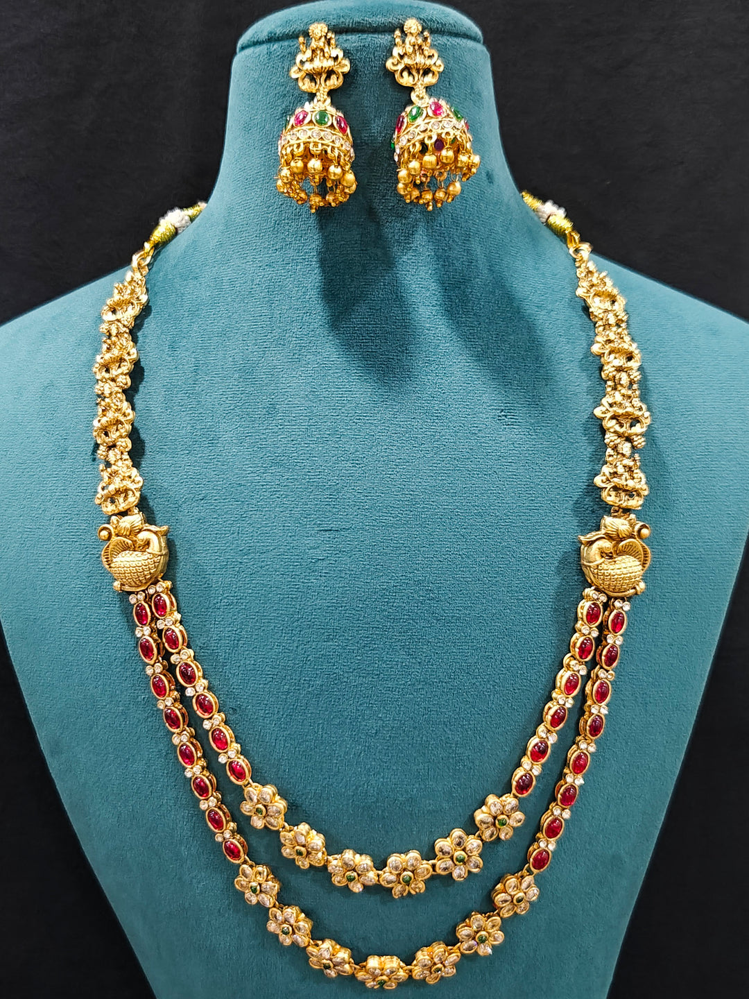 The Red & Green Karuna Temple Necklace Set featuring a traditional gold Long necklace and matching earrings with Red and green colored stones, Close-up of detailed temple carvings in artificial wedding jewelry.
