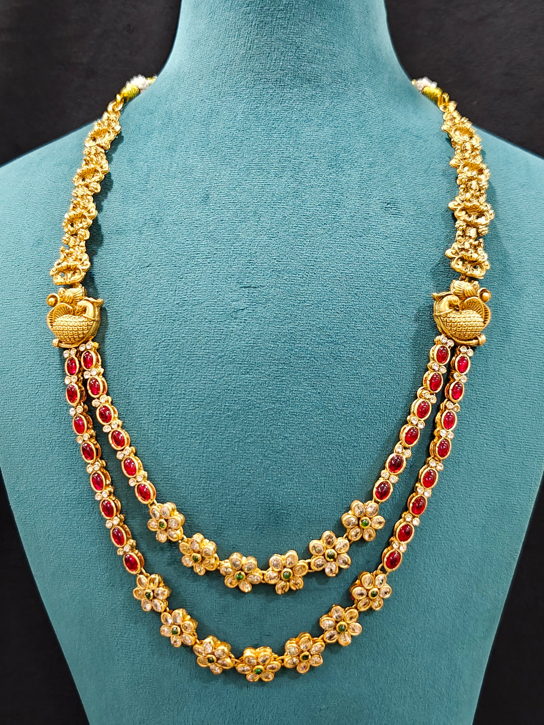 The Red & Green Karuna Temple Necklace Set featuring a traditional gold Long necklace and matching earrings with Red and green colored stones, Close-up of detailed temple carvings in artificial wedding jewelry.
