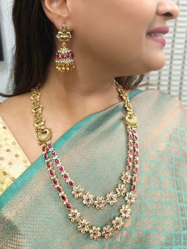 A woman wearing a green and gold saree with the Red Karuna Temple Necklace Set—featuring a double-layered gold necklace and matching jhumka earrings with red stones and floral motifs—smiling slightly, perfect for a wedding ceremony.