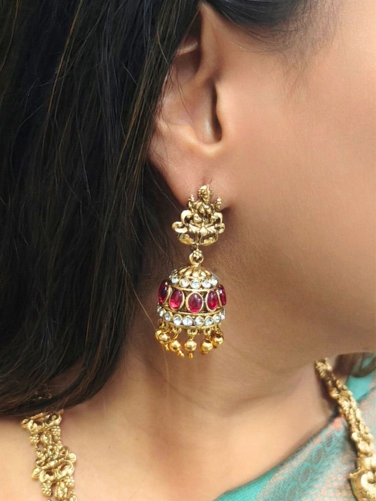 A woman wearing a green and gold saree with the Red Karuna Temple Necklace Set—featuring a double-layered gold necklace and matching jhumka earrings with red stones and floral motifs—smiling slightly, perfect for a wedding ceremony.