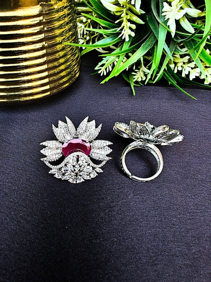 Two ornate, flower-shaped cocktail rings rest on a dark surface; one is the Red Megan Ring, featuring ruby red stones and sparkling American Diamond accents. The other ring is shown from behind, with green foliage and a gold object in the background.