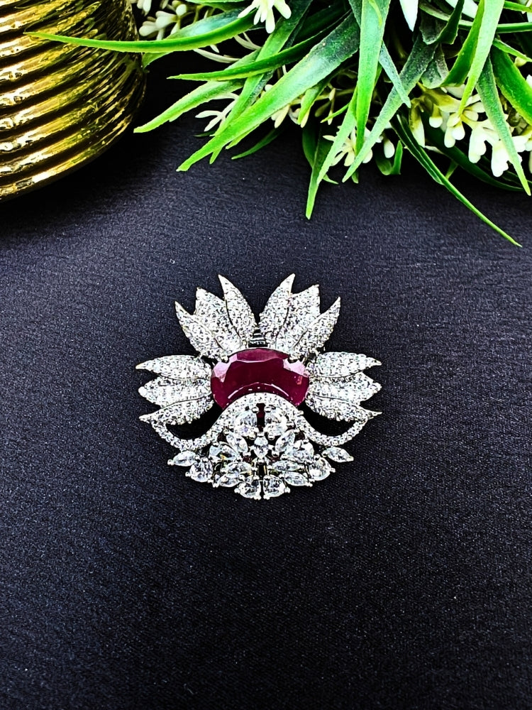 flower-shaped cocktail rings rest on a dark surface; this is the Red Megan Ring, featuring ruby red stones and sparkling American Diamond accents. The ring is shown with green foliage and a gold object in the background.