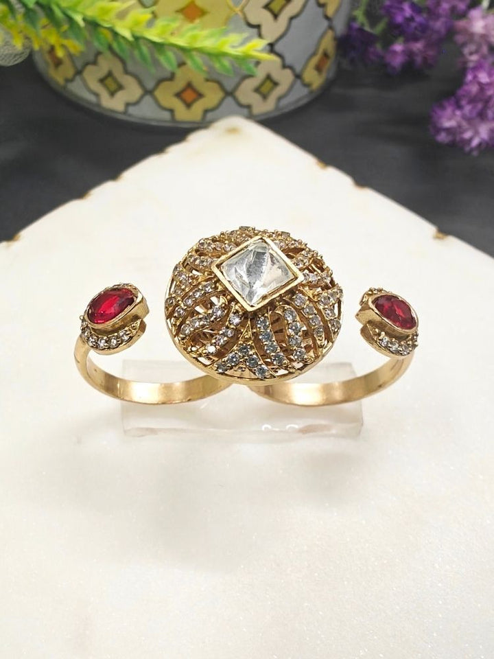 The Red Octavia Ring features a gold band with a round, ornate centerpiece set with a clear American Diamond and two smaller red gemstones on the side, displayed on a white surface with gold trim. Multi-finger ring, great for cocktail parties. 