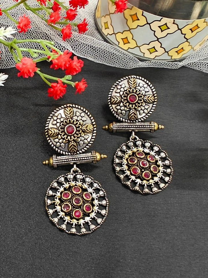 The Red Sheetal Earrings feature intricate circular brass designs with embedded American diamonds, offering an antique oxidized look. Displayed with red flowers and a patterned container, they’re perfect for a wedding ceremony. Perfect to wear on navratri. 