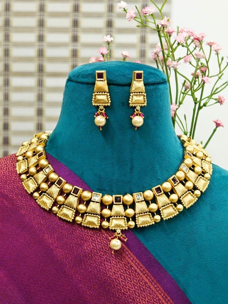 A mannequin displays the Red Sheetla Necklace Set, featuring a brass gold necklace and matching earrings with geometric patterns and pearl details, against a backdrop of pink flowers and a window.