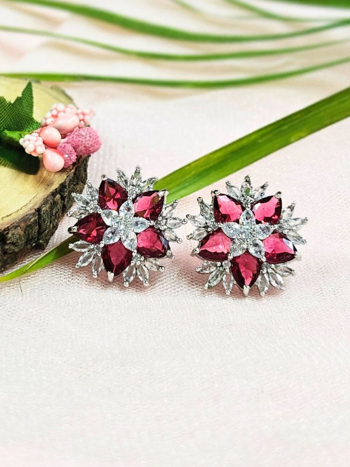 The Red Snowflake Earrings showcase Red colored stones along with American Diamonds in a starburst design, perched on a soft pink fabric with green leaves and a Pink flower.