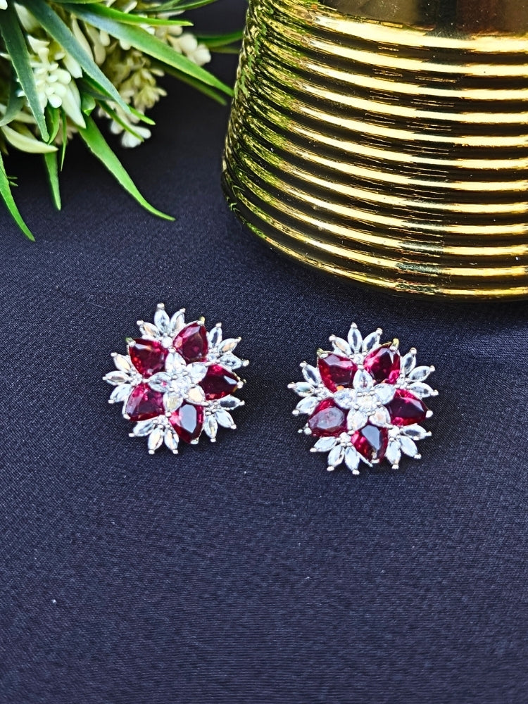 The Red Snowflake Earrings showcase Red colored stones along with American Diamonds in a starburst design, perched on a Black Surface slab with a gold-striped background.