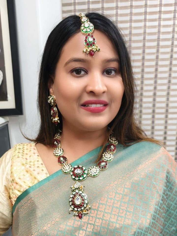 A woman smiles while looking to the side, wearing a green and gold saree with ethnic jewelry, including the Red Suhasini Necklace Set, earrings, and a kundan maang tikka, in front of a patterned background. The necklace is made with kundan detailing all around the necklace, with some colored stones, along with pearls hanging at the bottom. 