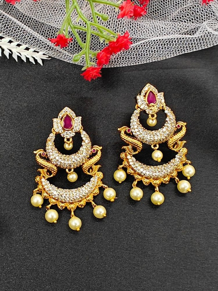 The Red Zarina Earrings are ornate gold and American Diamond earrings with pearl dangles and intricate detailing, displayed on a black surface with white netting and red flowers. Perfect to complement your wedding ceremony dress.