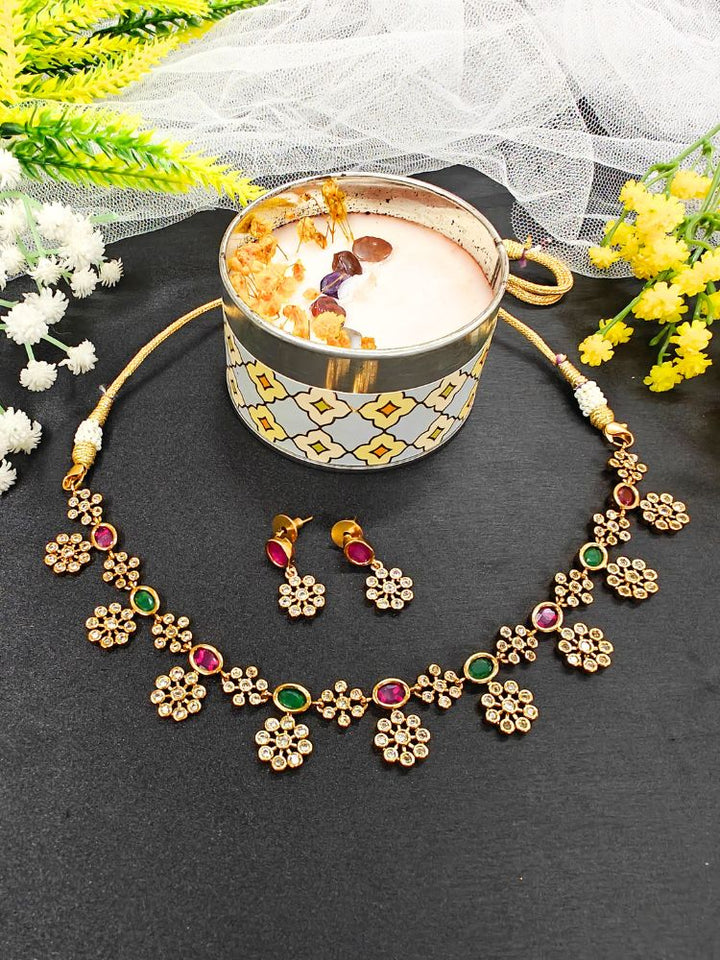 The Red and Green Rashi Necklace Set features a floral gold necklace with matching earrings, decorated with American Diamonds and stones, shown on a black surface beside a patterned cup of milk, surrounded by yellow and white flowers.