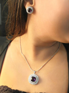 A woman wears the Renne Dark Purple Pendant Set by Peacoski—a silver necklace with a bold dark purple pendant and matching earrings, both accented with sparkling American Diamonds, ideal for complementing a cocktail gown.