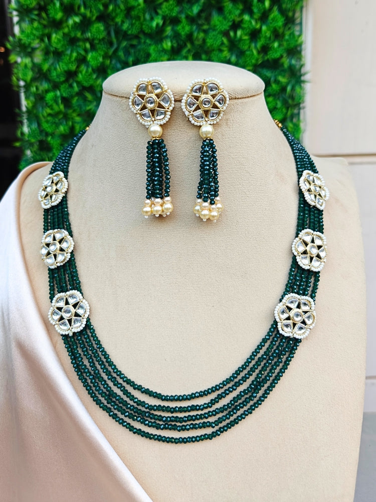 The Renuka Necklace Set features a multi-strand green crystal beaded necklace with five floral motifs, paired with matching ethnic earrings with pearl accents, displayed on a beige stand against a green backdrop.