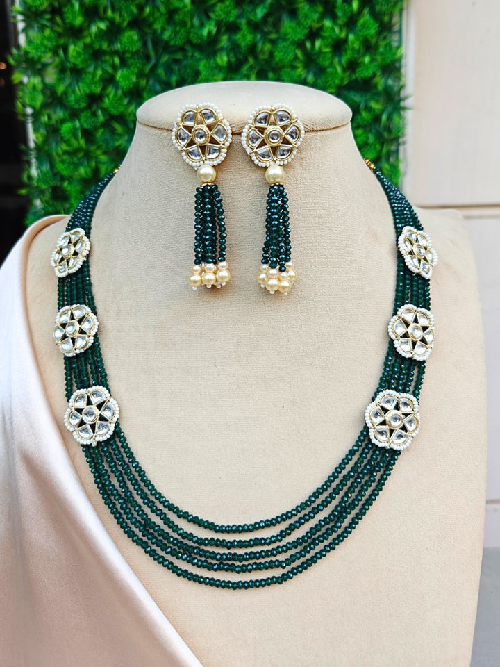 The Renuka Necklace Set features a multi-strand green crystal beaded necklace with five floral motifs, paired with matching ethnic earrings with pearl accents, displayed on a beige stand against a green backdrop.