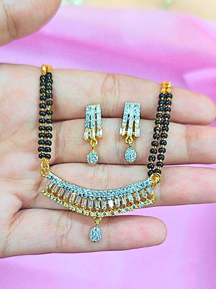 A hand holds the Ritu Mangalsutra—a gold necklace with black mangalsutra beads and matching earrings, pendant featuring sparkling American Diamonds—set against a soft pink background, perfect for your wedding ceremony dress. Diamond mangalsutra. Modern mangalsutra. 