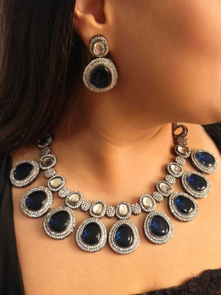 A woman with brown hair down smiles to the side, wearing the Royal Blue Elizabeth Necklace Set featuring a statement Victorian-style necklace and matching earrings with large colorful stones, paired with a sparkly dark top.