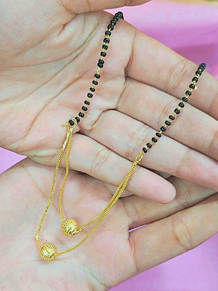 A hand holds the Rupali Mangalsutra—a gold necklace with black mangalsutra beads and matching earrings, pendant featuring sparkling American Diamonds—set against a soft pink background, perfect for your wedding ceremony dress. Diamond mangalsutra. Modern mangalsutra. 