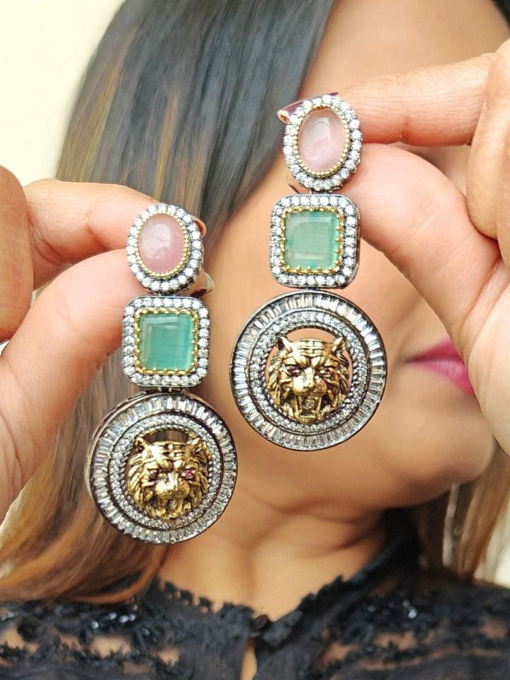 A woman in a black lace top holds up the Sabyasachi Inspired Earrings 1—Pink & Green earrings with gold lion head designs and sparkling American Diamonds, making them the perfect statement piece for a cocktail gown. Sabyasachi Earrings. American Diamond Earrings. Long Earrings. Party Earrings. 