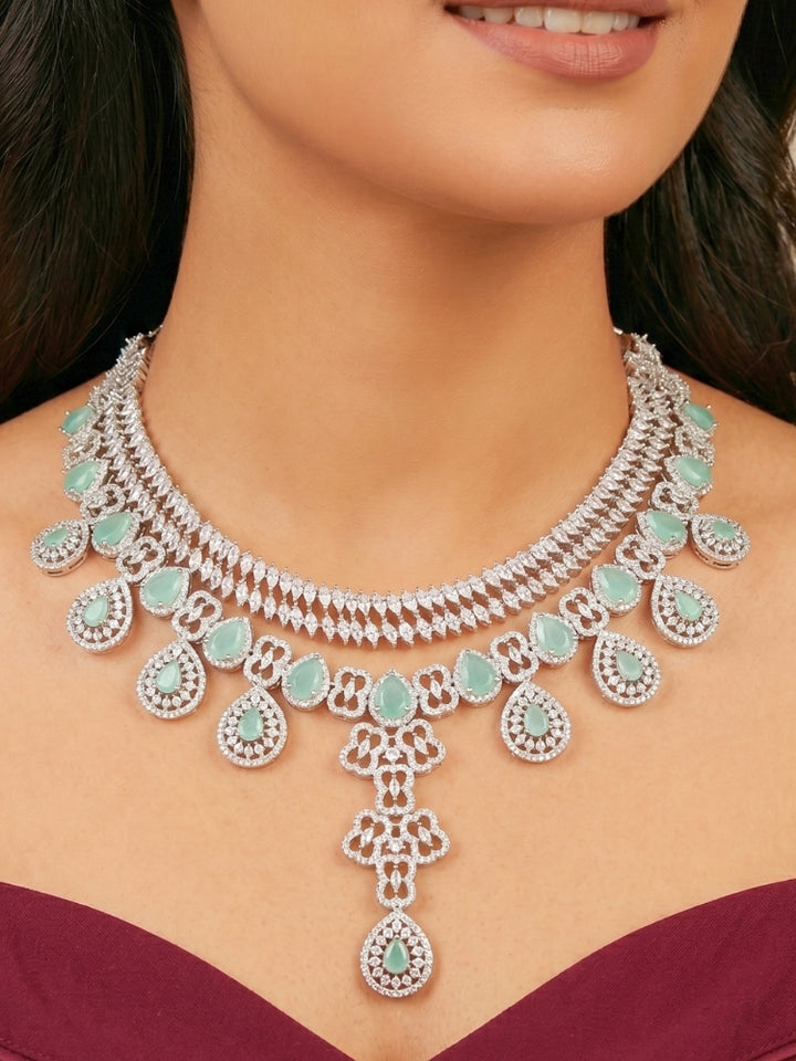 Close-up of a women wearing an ornate Western (Silver Polish) Collar Layered jewellery set and earrings with American Diamond and Mint Green Colored Stones. Reception-ready look featuring a statement American diamond jewelry set. Elegant wedding jewelry look featuring a premium artificial American diamond necklace. 