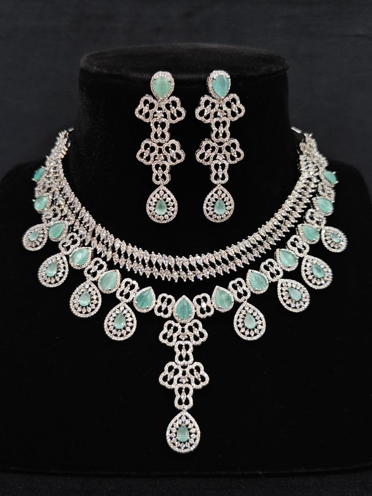Close-up of an ornate Western (Silver Polish) Collar Layered jewellery set and earrings with American Diamond and Mint Green Colored Stones. Reception-ready look featuring a statement American diamond jewelry set. Elegant wedding jewelry look featuring a premium artificial American diamond necklace.