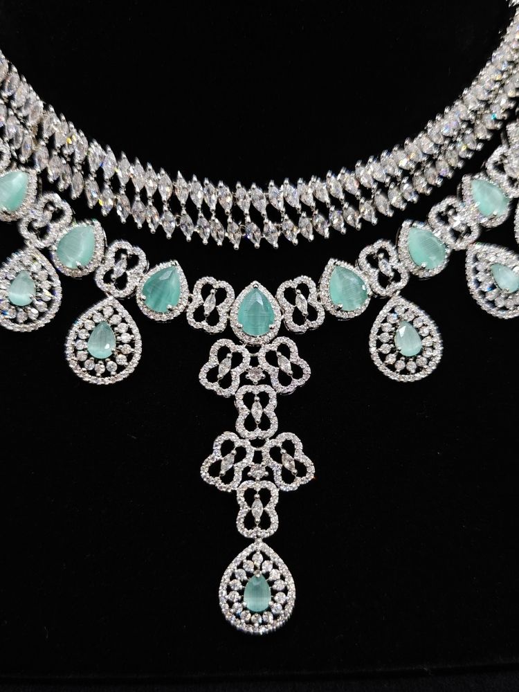 Close-up of an ornate Western (Silver Polish) Collar Layered jewellery set and earrings with American Diamond and Mint Green Colored Stones. Reception-ready look featuring a statement American diamond jewelry set. Elegant wedding jewelry look featuring a premium artificial American diamond necklace.