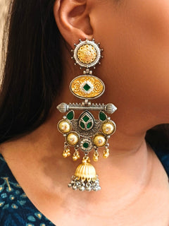 A close-up of a womans ear wearing Sanjukta Green Earrings, featuring ornate multicolored designs, American Diamond accents, gold and maroon details, intricate patterns, dangling beads, and a bell-shaped charm at the bottom. These are oxidized german silver earrings perfect for navratri earrings. 