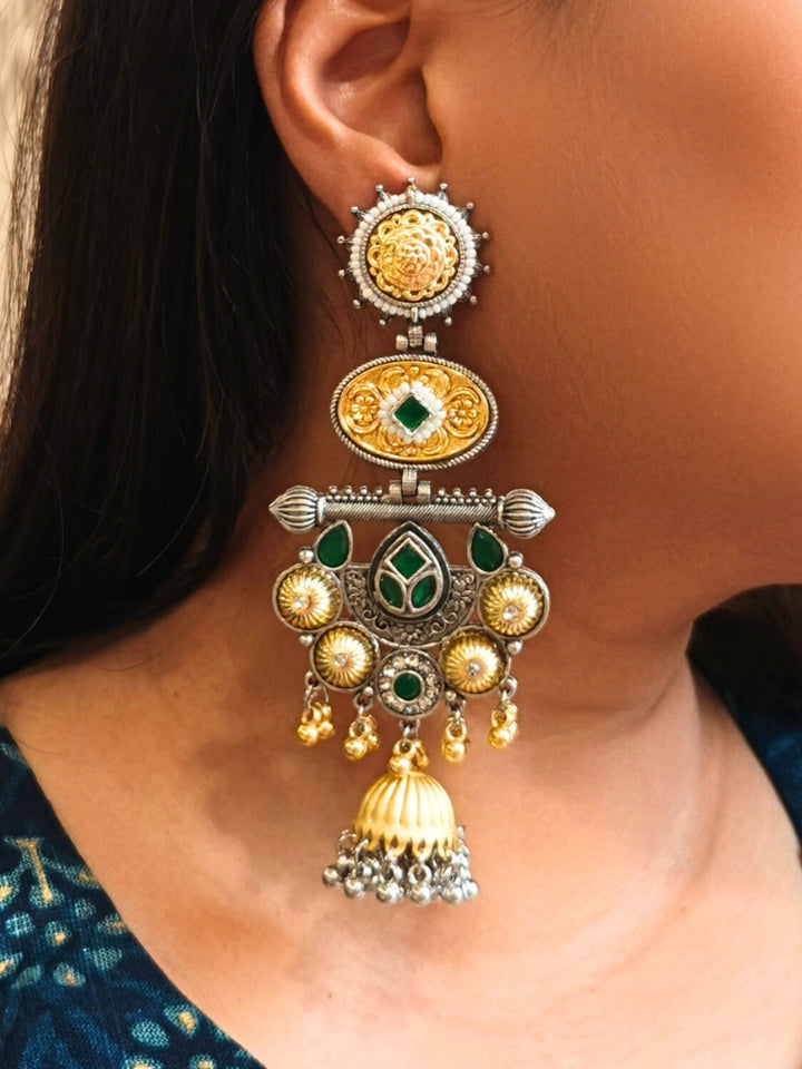 A close-up of a womans ear wearing Sanjukta Green Earrings, featuring ornate multicolored designs, American Diamond accents, gold and maroon details, intricate patterns, dangling beads, and a bell-shaped charm at the bottom. These are oxidized german silver earrings perfect for navratri earrings. 