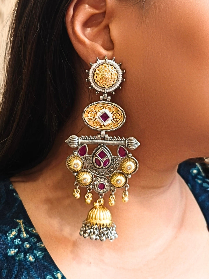 A close-up of a womans ear wearing Sanjukta Pink Earrings, featuring ornate multicolored designs, American Diamond accents, gold and maroon details, intricate patterns, dangling beads, and a bell-shaped charm at the bottom. These are oxidized german silver earrings perfect for navratri earrings. 
