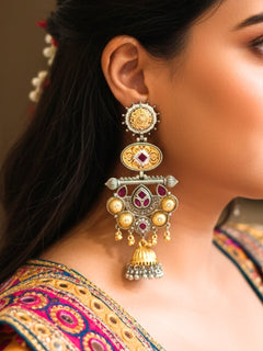 A close-up of a womans ear wearing Sanjukta Pink Earrings, featuring ornate multicolored designs, American Diamond accents, gold and maroon details, intricate patterns, dangling beads, and a bell-shaped charm at the bottom. These are oxidized german silver earrings perfect for navratri earrings. 
