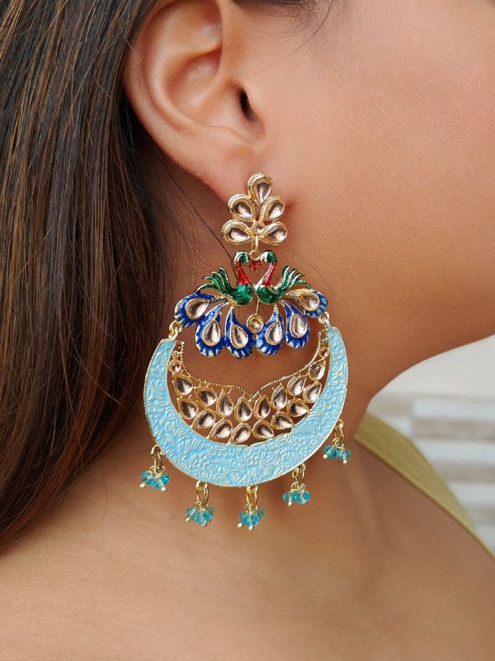 A close-up of a womans ear wearing Sarika Blue Earrings, featuring large, ornate peacock designs with gold, red, green, and blue detailing, embellished with Kundan and pearls and finished with small dangling red beads.