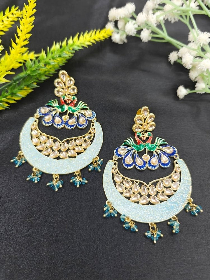 Sarika Blue Earrings, featuring large, ornate peacock designs with gold, blue, green, and red detailing, embellished with Kundan and pearls and finished with small dangling red beads, shown on a black background. 