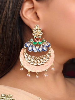 A close-up of a womans ear wearing Sarika Peach Earrings, featuring large, ornate peacock designs with gold, red, green, and blue detailing, embellished with Kundan and pearls and finished with small dangling red beads.