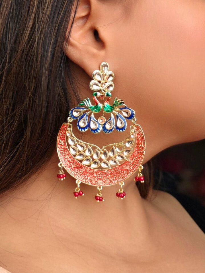 A close-up of a womans ear wearing Sarika Red Earrings, featuring large, ornate peacock designs with gold, red, green, and blue detailing, embellished with Kundan and pearls and finished with small dangling red beads.