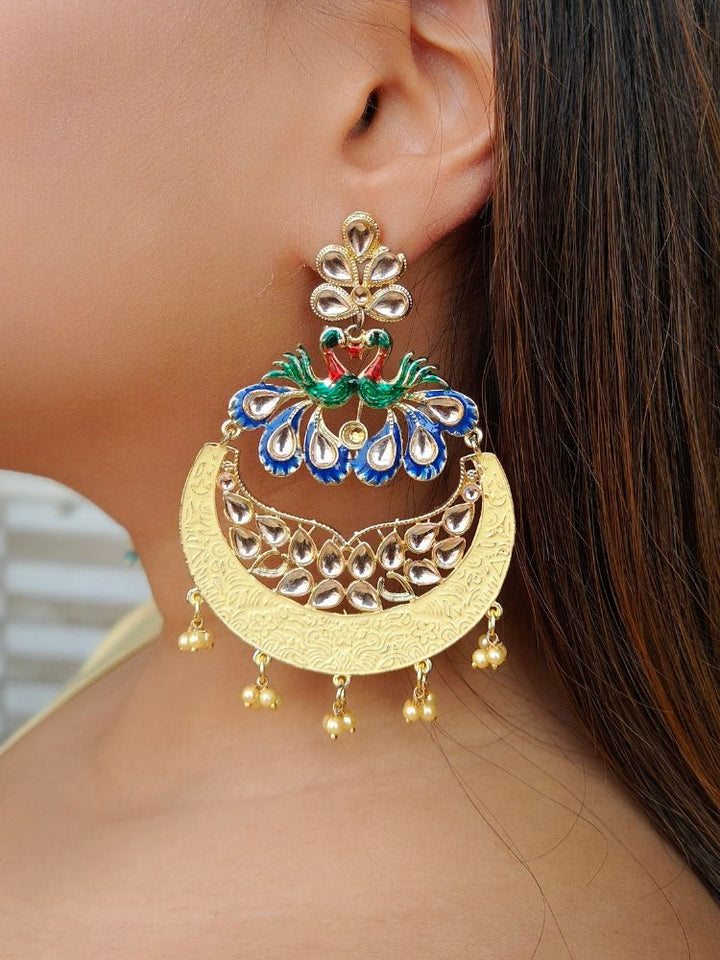 A close-up of a womans ear wearing Sarika Yellow Earrings, featuring large, ornate peacock designs with gold, red, green, and blue detailing, embellished with Kundan and pearls and finished with small dangling red beads.