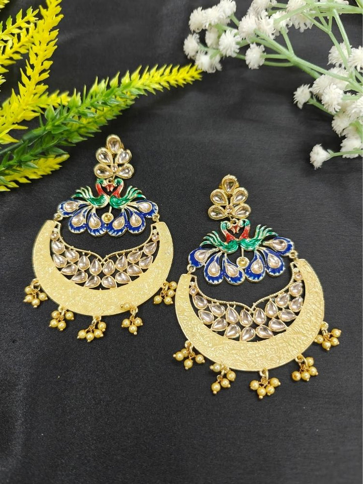 Sarika Yellow Earrings, featuring large, ornate peacock designs with gold, red, green, and blue detailing, embellished with Kundan and pearls and finished with small dangling red beads, shown on a black background with green and white flowers on the back. 