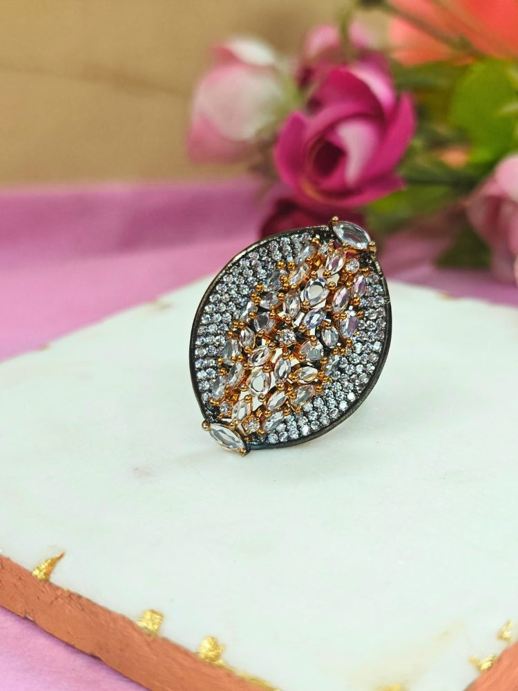 The Sasha Ring, featuring intricate gold and silver detailing with jeweled accents, is displayed on a white marble slab. Soft pink and red artificial roses blur in the background—a perfect American Diamond cocktail ring for your wedding ceremony dress.