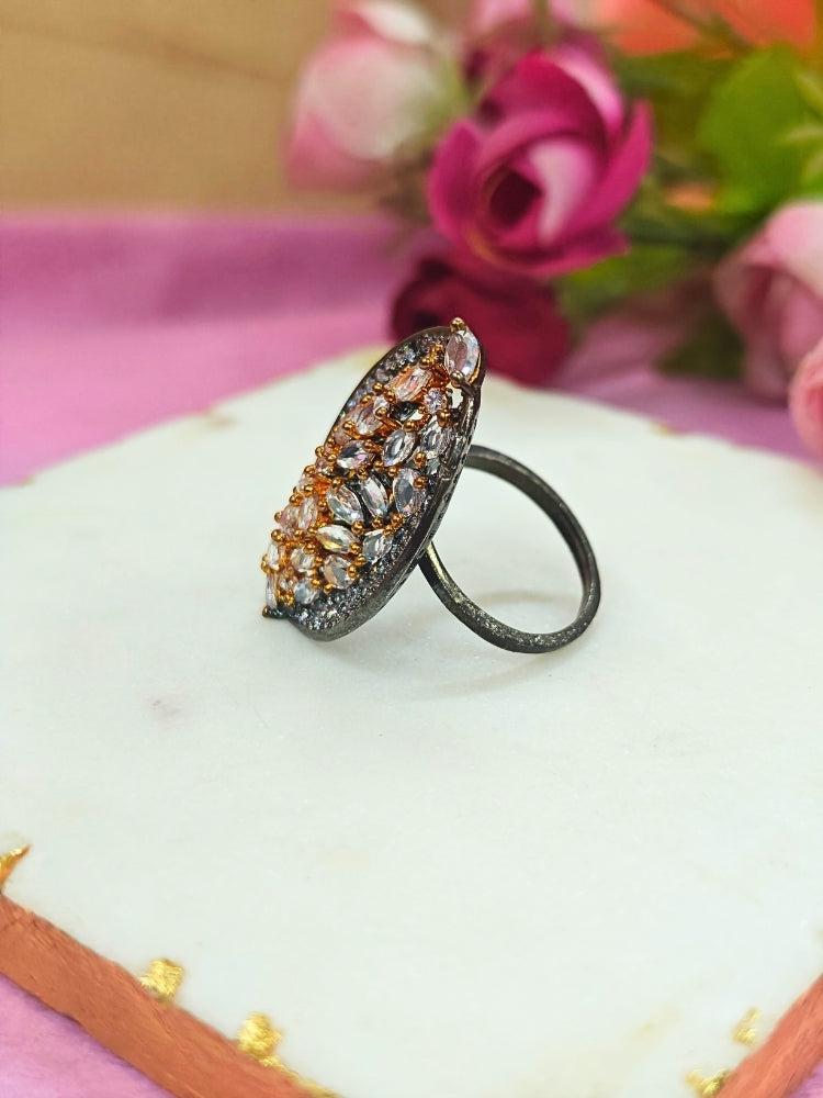 The Sasha Ring, featuring intricate gold and silver detailing with jeweled accents, is displayed on a white marble slab. Soft pink and red artificial roses blur in the background—a perfect American Diamond cocktail ring for your wedding ceremony dress.