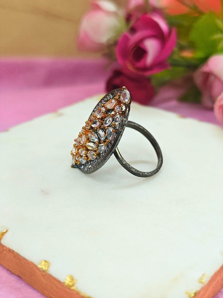 The Sasha Ring, featuring intricate gold and silver detailing with jeweled accents, is displayed on a white marble slab. Soft pink and red artificial roses blur in the background—a perfect American Diamond cocktail ring for your wedding ceremony dress.