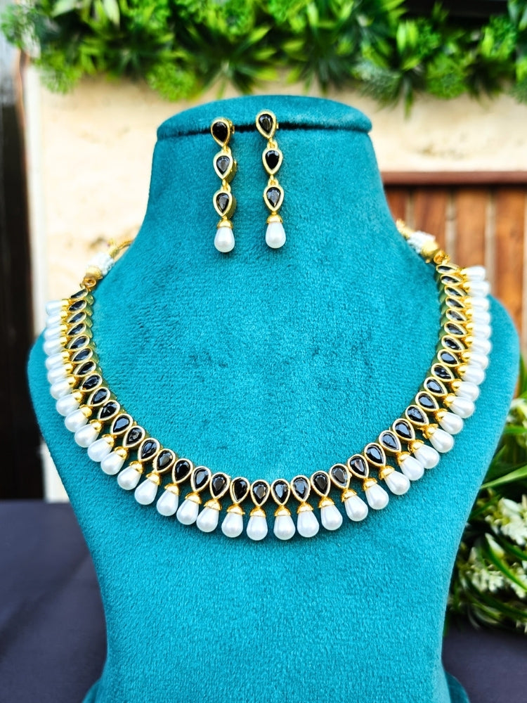 The Saumya Necklace Set features a gold necklace and matching earrings with teardrop black stones,  each stone attached to high quality pearls, elegantly displayed on teal velvet—ideal for complementing your wedding ceremony dress.