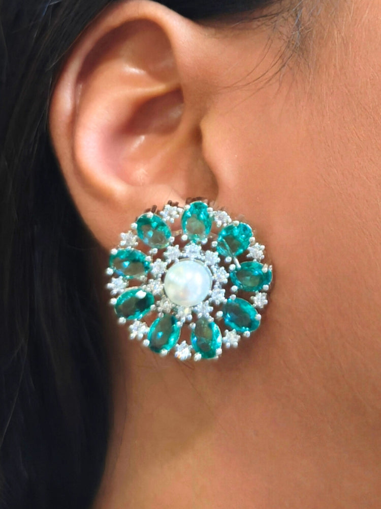 A close-up of a womans ear wearing the Sea Green Blossom stud Earrings—large, circular Sea Green earrings with a pearl at the center, colorful accents, and a border of sparkling American Diamonds. The stud earrings give a flower earrings feel. 
