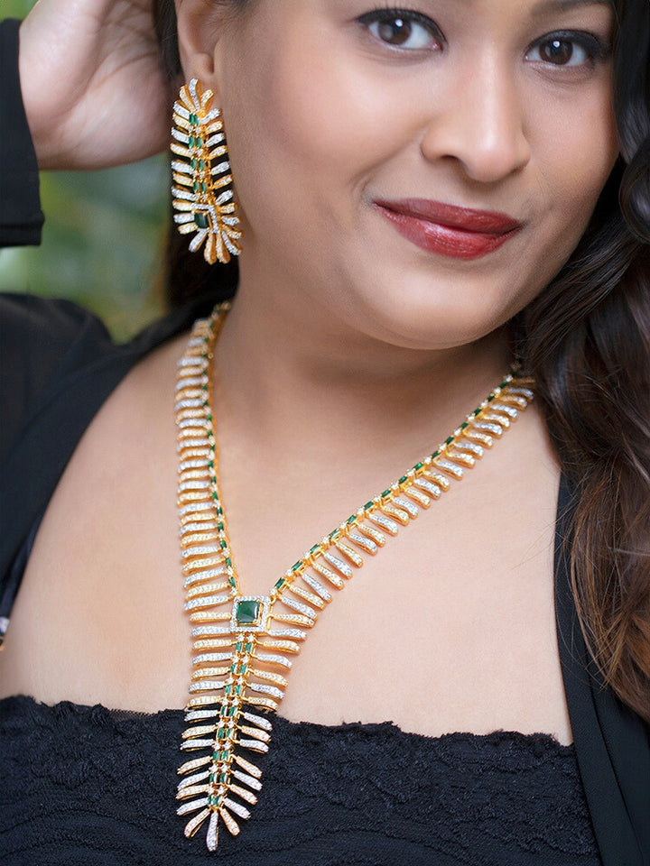 A woman in a strapless black dress showcases the Selvaggia long Necklace Set, featuring sparkling American Diamonds and white stones, with large earrings and an intricate green necklace adorned with gemstone accents.