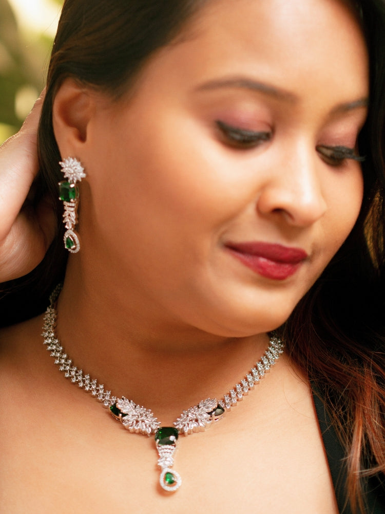 A woman wears the Serena Green Necklace Set—featuring a striking green necklace and matching earrings with colored stones and sparkling American diamonds—against a blurred background. American Diamond Necklace with Green colored stones. 