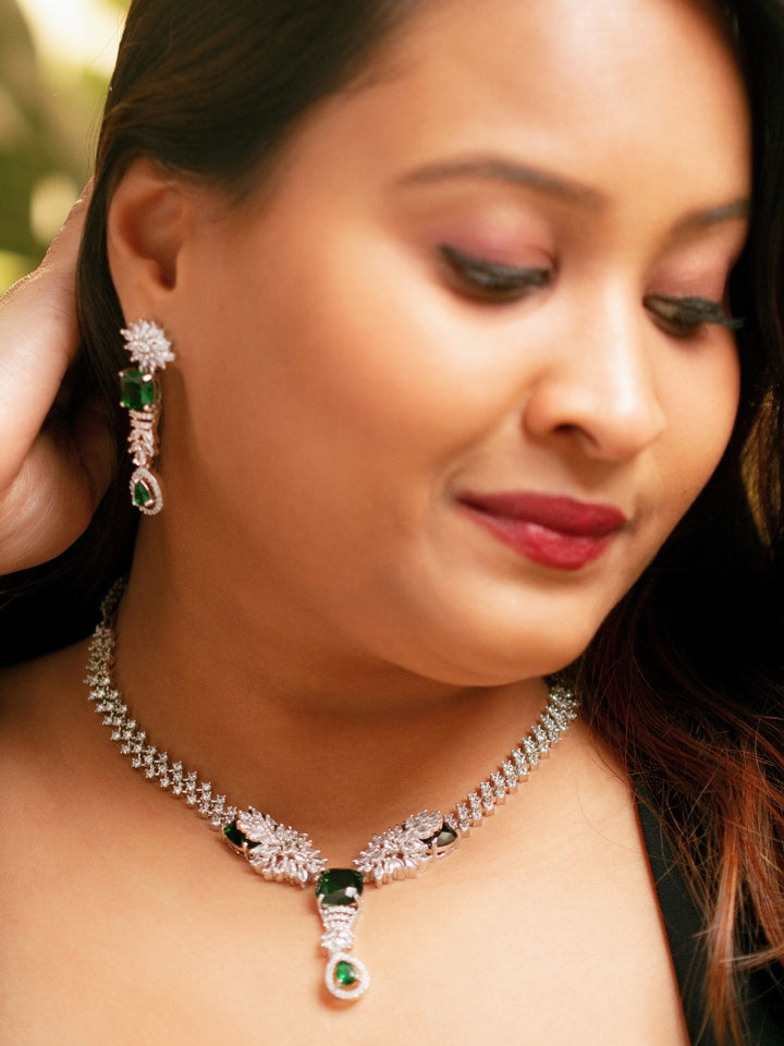 A woman wears the Serena Green Necklace Set—featuring a striking green necklace and matching earrings with colored stones and sparkling American diamonds—against a blurred background. American Diamond Necklace with Green colored stones. 
