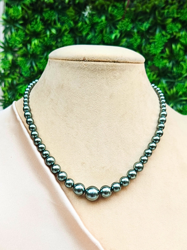 A mannequin in teal velvet showcases the Sharon Olive Green Necklace Set—a single-strand Olive Green pearl necklace—elegantly draped with gold and beige embroidery on one shoulder, against a softly patterned background. Ideal for wedding ceremonies.