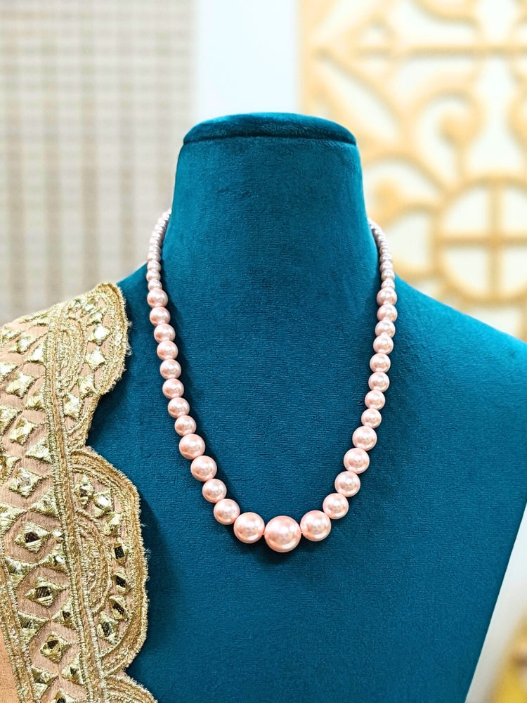A mannequin in teal velvet showcases the Sharon Peach Necklace Set—a single-strand peach pearl necklace—elegantly draped with gold and beige embroidery on one shoulder, against a softly patterned background. Ideal for wedding ceremonies.