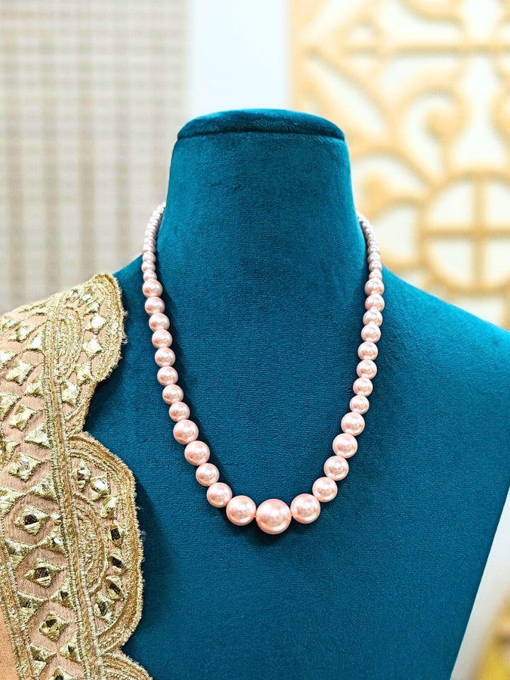 A mannequin in teal velvet showcases the Sharon Peach Necklace Set—a single-strand peach pearl necklace—elegantly draped with gold and beige embroidery on one shoulder, against a softly patterned background. Ideal for wedding ceremonies.