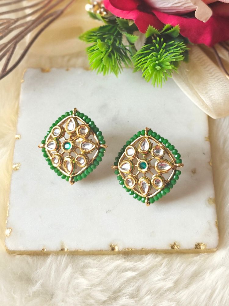 Sharvisha Dark Green Earrings: Square-shaped Kundan earrings with teardrop white Kundan accents, surrounded by Dark Green crystal stones, displayed on a marble-like surface with flowers in the background.
