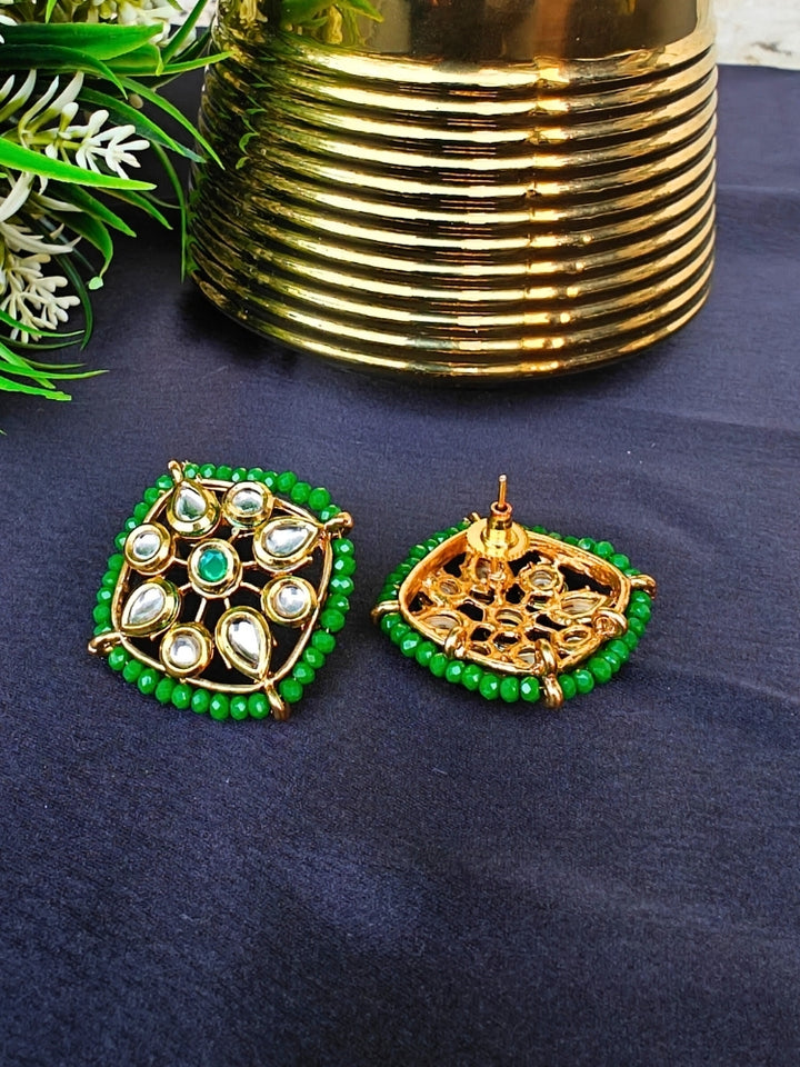 Sharvisha Dark Green Earrings: Square-shaped Kundan earrings with teardrop white Kundan accents, surrounded by Dark Green crystal stones, displayed on a Black surface with flowers & Leaves in the background.