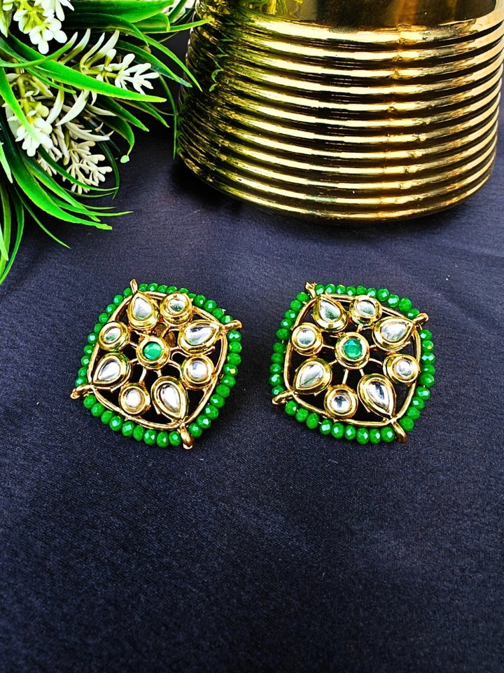 Sharvisha Dark Green Earrings: Square-shaped Kundan earrings with teardrop white Kundan accents, surrounded by Dark Green crystal stones, displayed on a Black surface with flowers & Leaves in the background.