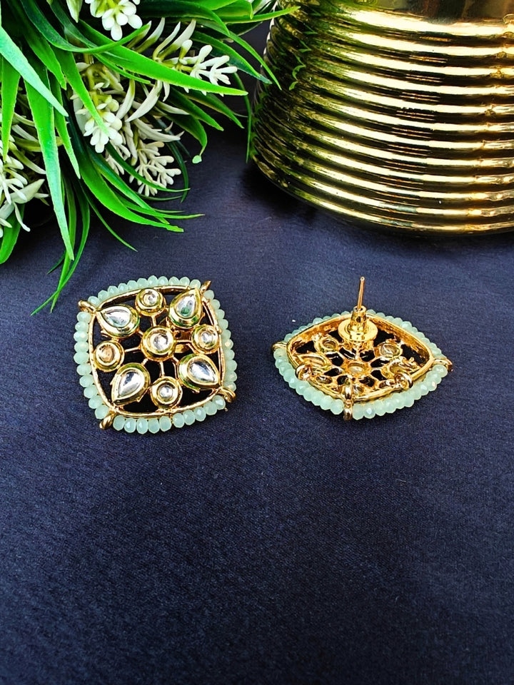 Sharvisha Mint Green Earrings: Square-shaped Kundan earrings with teardrop white Kundan accents, surrounded by Mint Green crystal stones, displayed on a Black surface with flowers & Leaves in the background.